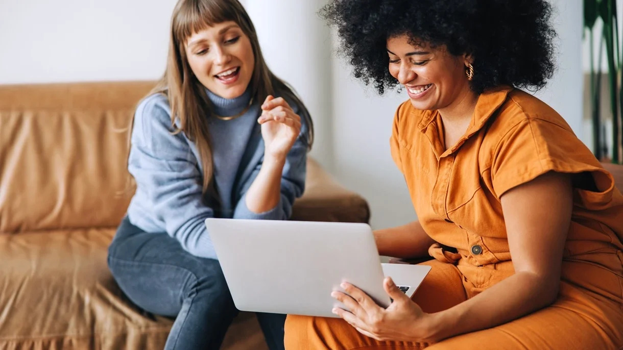 Two women seated on a couch, engaged with a laptop, sharing ideas and collaborating in a comfortable setting.