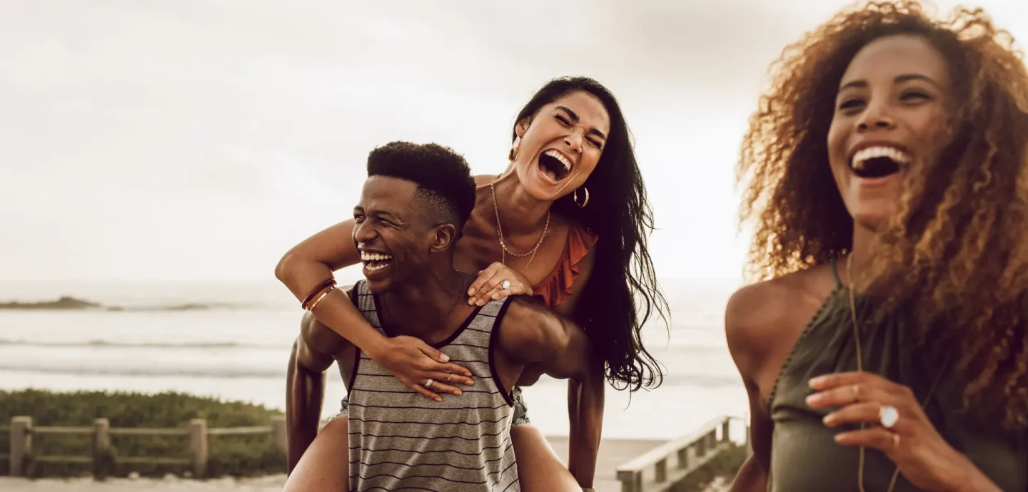 Three young people joyfully laughing and smiling together on a sunny beach, enjoying their time by the ocean.