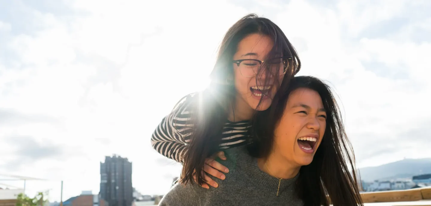 Two women joyfully laughing and smiling together on a rooftop, enjoying a sunny day and each other's company.